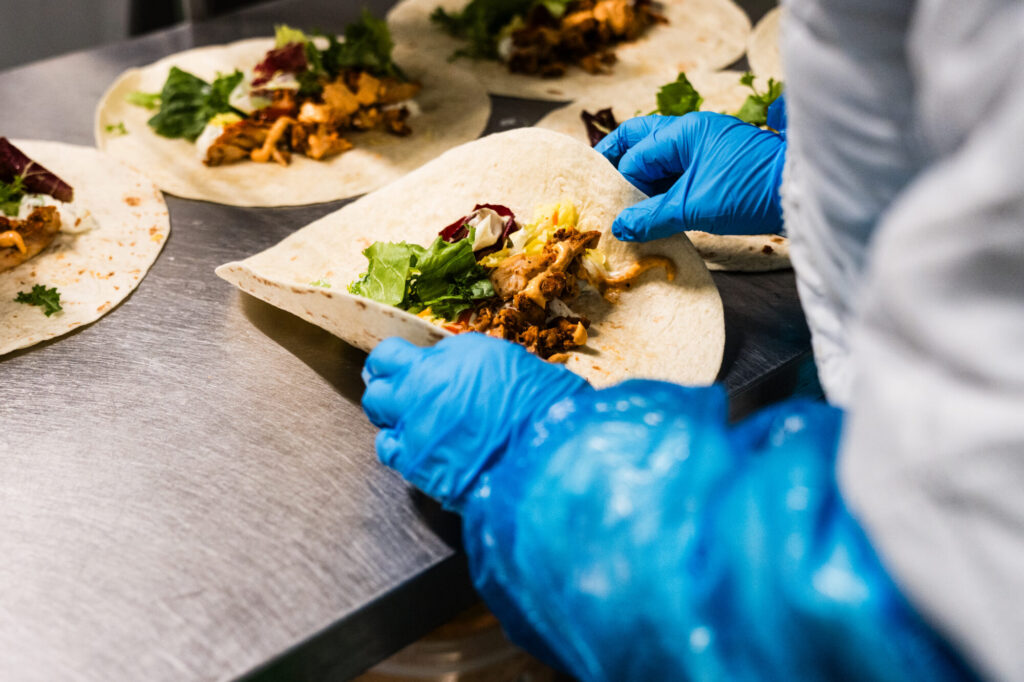 Hände in Handschuhen belegen eine Tortilla mit Salat und Hähnchen